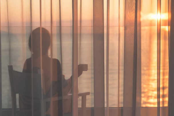 Naked beautiful female wrapped in a blanket sitting on a chair with cup of coffee behind the transparent curtains on the glass balcony early morning at sunrise. Sea view - Φωτογραφία, εικόνα