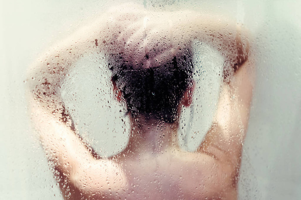 濡れたミストガラスの後ろにシャワーで髪を洗う裸の美しい女性のシルエット. - 写真・画像