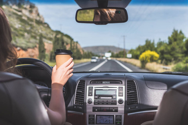 The girl is driving on the highway and holding paper cup of coffee . View from the back seat of the car  - Foto, imagen