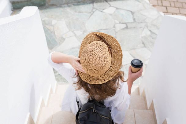 жінка з рюкзаком в солом'яному капелюсі і паперовою чашкою кави сидить на сходах і відпочиває в курортному місті. Індивідуальний мандрівник
. - Фото, зображення