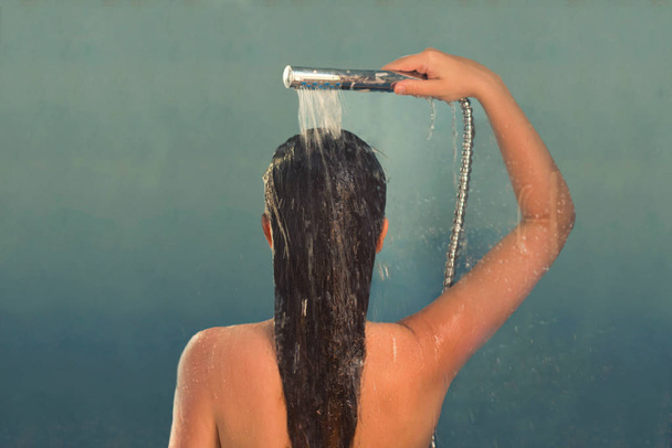 Back view of beautiful naked young woman taking shower in shower cabin. Drops on the transparent glass door - 写真・画像