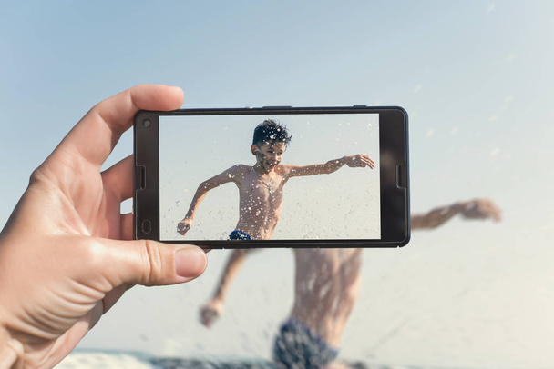Žena, která si na telefonu fotografovala chlapce na pláži. Šťastný mladík se baví na písku na pláži. Cestovní a rodinná koncepce - Fotografie, Obrázek