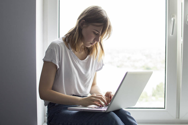 jong meisje zitten in de buurt van het raam met een laptop, ze maakt gebruik van de computer thuis op de vensterbank, een vrouwelijke freelance, kopieer ruimte voor tekst - Foto, afbeelding