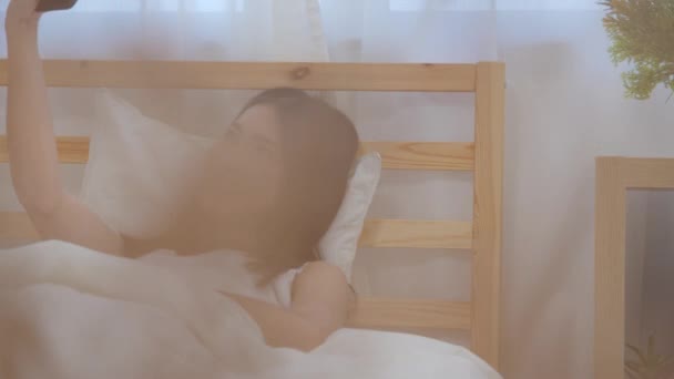 Pomalý pohyb-mladá asijská žena, která používá smartphone a kontroluje společenské sdělovací prostředky, se šťastně usmívá, když leží na posteli, když se ráno probudila, atraktivní Japonka se usmívá v ložnici doma. - Záběry, video