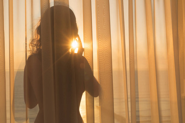 Naked beautiful woman in the headphones stands behind the transparent curtain on the balcony at sunrise on the tropical resort - Foto, immagini