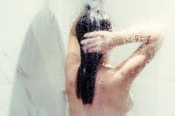 Silhouette of naked beautiful woman washing hair in shower behind wet misted glass. - Foto, immagini
