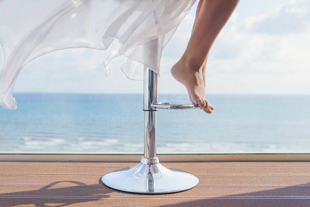 woman is sitting on a bar stool in a summer cafe on the sea and sky background. Bottom view on barefoot female legs - Foto, Imagem