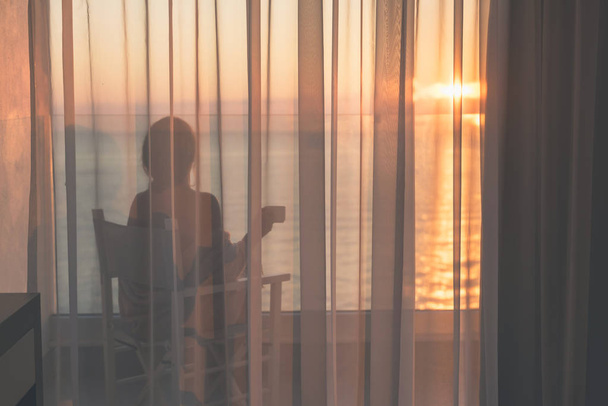 Çıplak güzel kadın gün doğumunda cam balkonda şeffaf perdeler arkasında kahve fincan ı sehpa ile bir sandalyede oturan bir battaniye sarılmış. Deniz manzarası - Fotoğraf, Görsel