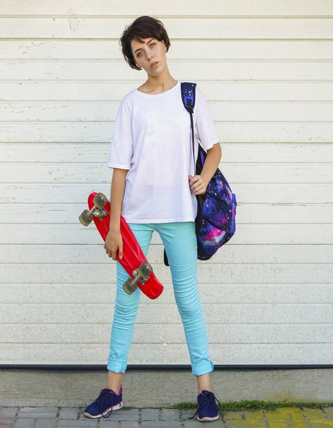 Fashion summer hipster cool woman in white t-shirt and colorful trousers with skateboard outdoors. garage doors on background. full-length - Photo, Image