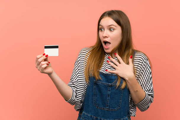 Jeune femme avec salopettes sur le mur rose tenant une carte de crédit et surpris
 - Photo, image