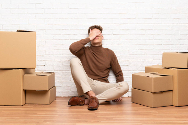 Handsome young man moving in new home among boxes covering eyes by hands - Photo, Image
