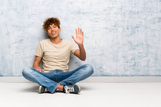 Молодий афроамериканець чоловік сидить на підлозі вітання з рукою з щасливим виразом - Фото, зображення