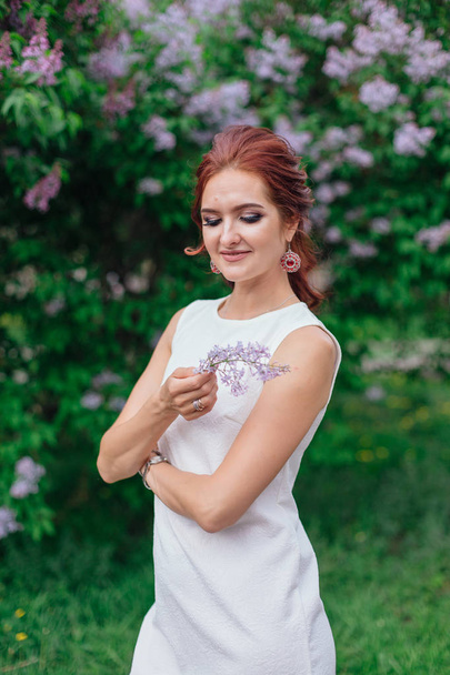 Charming woman wearing beautiful white dress standing next to lilac bush - Zdjęcie, obraz