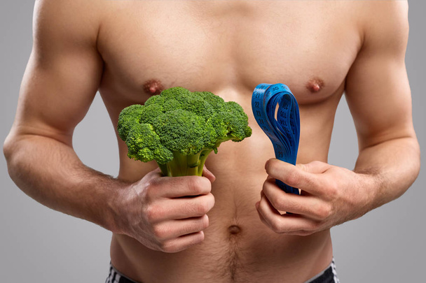 Crop muscular guy with broccoli and tape measure - Фото, изображение