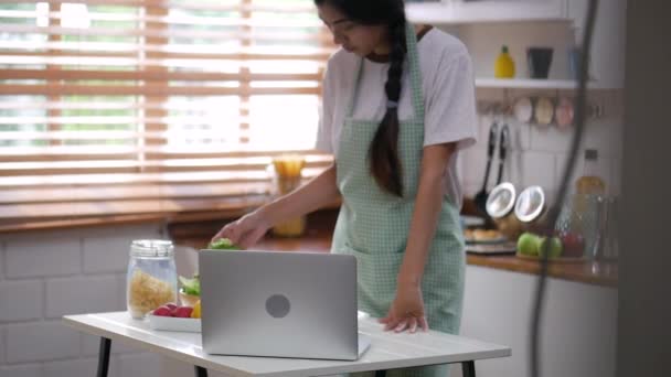 Young asian woman using laptop and ingredients for making salad, vegetable, strawberry cooking a healthy breakfast in kitchen at home. - Séquence, vidéo