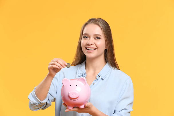 Young woman putting coin into piggy bank on color background - Zdjęcie, obraz