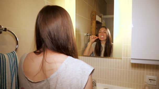 Η νεαρή γυναίκα βουρτσίζει τα δόντια της στο μπάνιο το πρωί κοιτάζοντας καθρέφτη. - Πλάνα, βίντεο