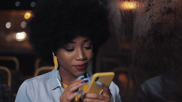 Attractive young african american woman using smartphone riding at public transport and looking out the window. Night time. City lights background. - Felvétel, videó