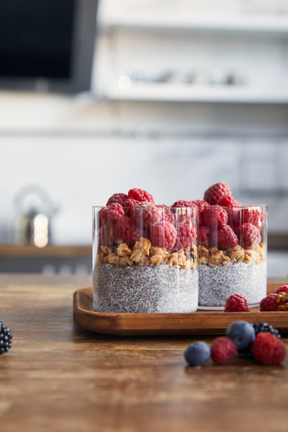 selective focus of yogurt with chia seeds, oat flakes and raspberries on wooden tray in kitchen - Photo, Image