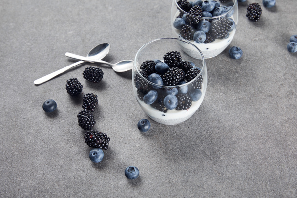 tasty yogurt with chia seeds, blueberries and blackberries in glasses near teaspoons and scattered berries on marble surface  - 写真・画像