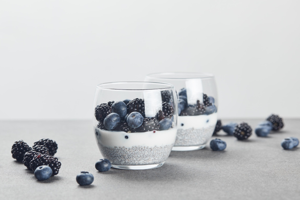 selektiver Fokus von leckerem Joghurt mit Chiasamen, Blaubeeren und Brombeeren in Gläsern nahe verstreuter Beeren auf Marmoroberfläche isoliert auf grau - Foto, Bild