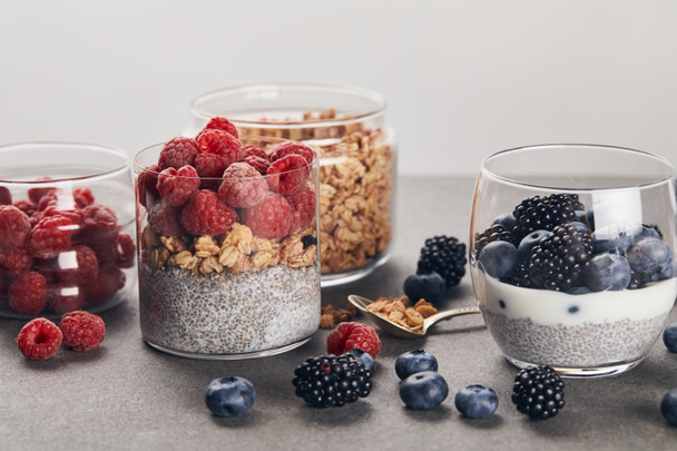 glasses with yogurt, berries and oat flakes near scattered berries on marble surface isolated on grey  - Foto, afbeelding