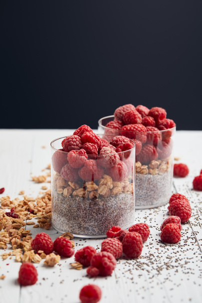 selective focus of glasses with yogurt, oat flakes and raspberries near scattered chia seeds, oat flakes and berries on white wooden table isolated on black - Foto, afbeelding