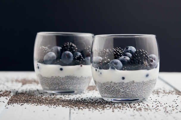 selective focus of yogurt with chia seeds, blueberries, blackberries near scattered seeds on white wooden isolated on black - Foto, Imagen