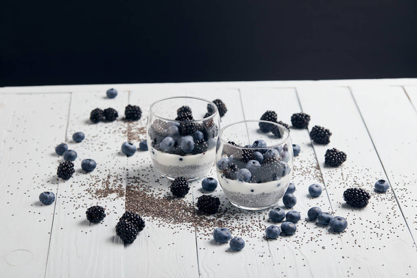 tasty yogurt with chia seeds, blueberries, blackberries near scattered seeds and berries on white wooden isolated on black - Fotoğraf, Görsel
