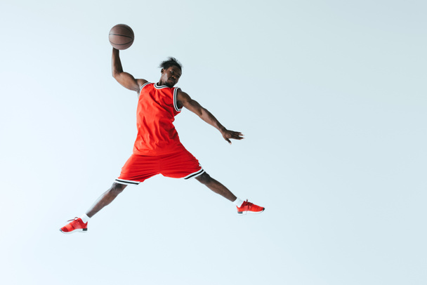 灰色で孤立したバスケットボールをしながら、アフリカ系アメリカ人のスポーツマンがジャンプ - 写真・画像