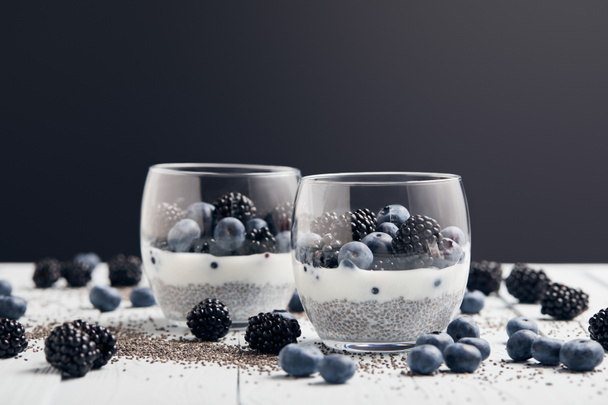 selective focus of yogurt with chia seeds and berries in glasses near scattered seeds, blueberries and blackberries on white table isolated on black - Photo, Image