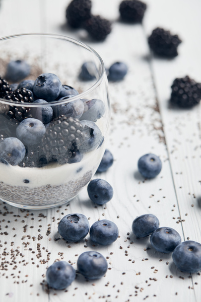 selective focus of blackberries, blueberries and yogurt in glass near scattered chia seeds and berries on white wooden table - Фото, изображение