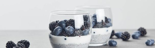 shot panorámico de sabroso yogur con semillas de chía, arándanos y moras en vasos cerca de bayas dispersas en la superficie de mármol aislado en gris
 - Foto, Imagen