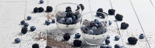 panoramic shot of tasty yogurt with chia seeds, blueberries, blackberries near scattered seeds and berries on white wooden isolated on black - Photo, Image