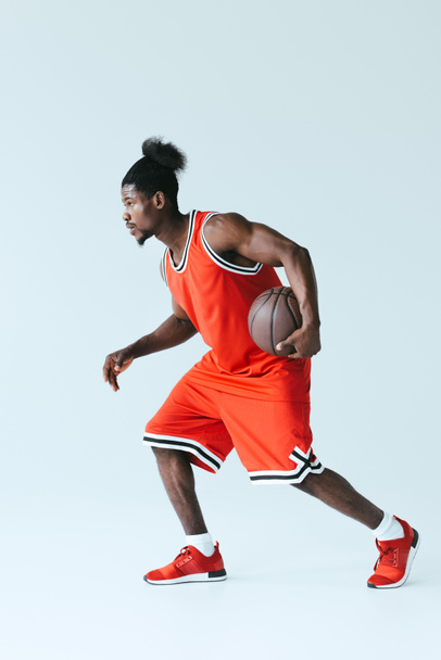 灰色の背景にバスケットボールをする赤いスポーツウェアのアフリカ系アメリカ人スポーツマン - 写真・画像