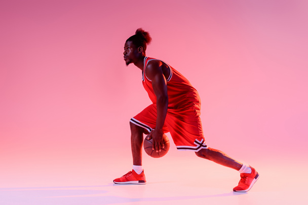 グラデーションと照明でピンクの背景にバスケットボールをプレイハンサムなアフリカ系アメリカ人のスポーツマン - 写真・画像