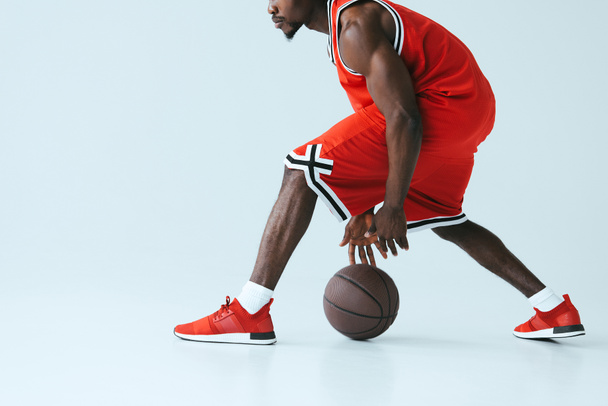灰色の背景にバスケットボールをプレイ赤いスポーツウェアでアフリカ系アメリカ人のスポーツマンのトリミングビュー - 写真・画像