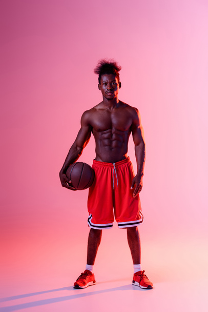 ημίγυμνος Αφρικανός Αμερικανός παίκτης μπάσκετ κοιτάζοντας την κάμερα σε ροζ και μωβ φόντο ντεγκραντέ με φωτισμό - Φωτογραφία, εικόνα