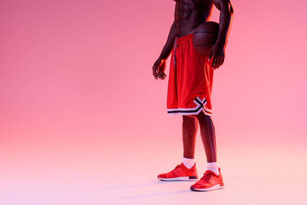 グラデーションと照明でピンクの背景にボールを保持するシャツのないアフリカ系アメリカ人バスケットボール選手のトリミングビュー - 写真・画像