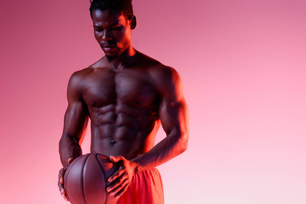 グラデーション付きピンクの背景にボールを保持する筋肉の胴体を持つ深刻なアフリカ系アメリカ人のスポーツマン - 写真・画像