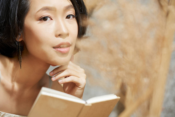 Portrait of attractive young Vietnamese woman with flawless skin holding small book looking at camera - Foto, Imagem