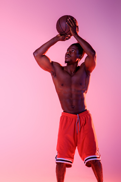 handsome, muscular african american sportsman playing basketball on pink and purple gradient background - Fotografie, Obrázek
