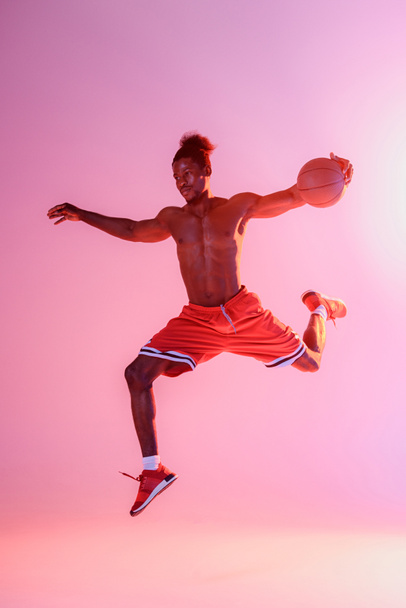 ピンクと紫のグラデーションの背景にバスケットボールをしながらジャンプ赤いショートパンツでシャツのないアフリカ系アメリカ人のスポーツマン - 写真・画像