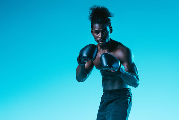 Selbstbewusster afrikanisch-amerikanischer Boxer mit muskulösem Oberkörper blickt in die Kamera auf blauem Hintergrund - Foto, Bild