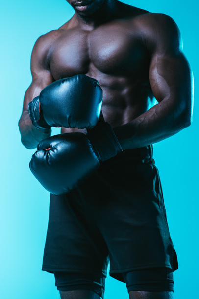 partial view of shirtless, muscular african american sportsman in boxing gloves on blue background - Foto, imagen