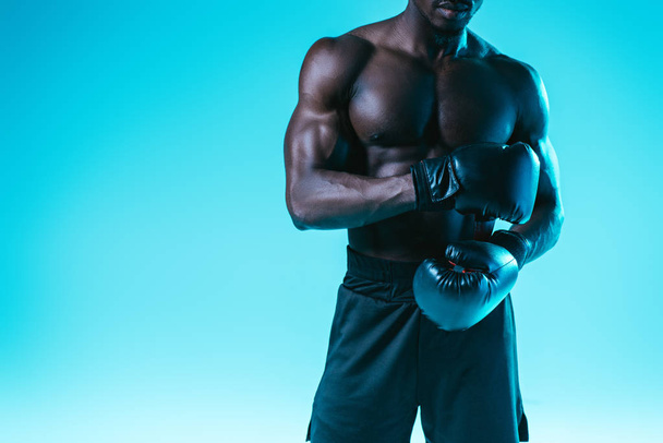 Ausschnittansicht eines afrikanisch-amerikanischen Boxers mit muskulösem Oberkörper, der auf blauem Hintergrund posiert - Foto, Bild