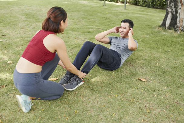 Fiatal nő gazdaság lábát a barátja, aki csinál crunches a földön a parkban - Fotó, kép