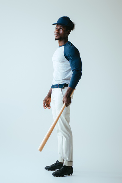 handsome african american sportsman looking away while holding baseball bat on grey background - 写真・画像