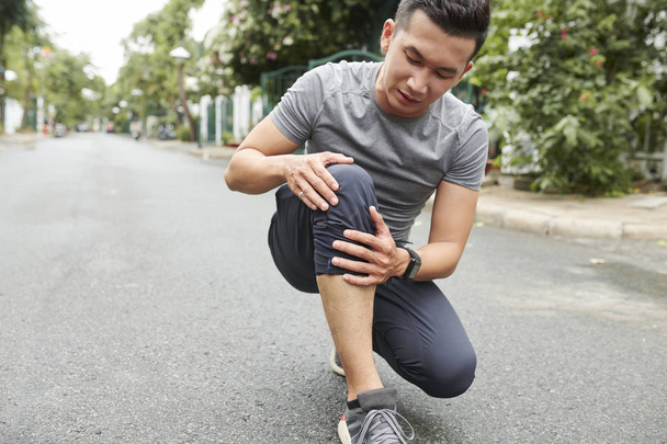 Young Vietnamese sportsman suffering from pain in knee after intense training outdoors - Fotó, kép