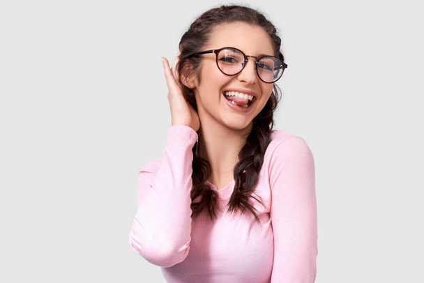 Joyful young woman showing her tongue, blinking with an eye and wearing round trendy transparent eyewear and long sleeve shirt while posing on white studio wall. Positive smiling female making grimace - Foto, Imagem